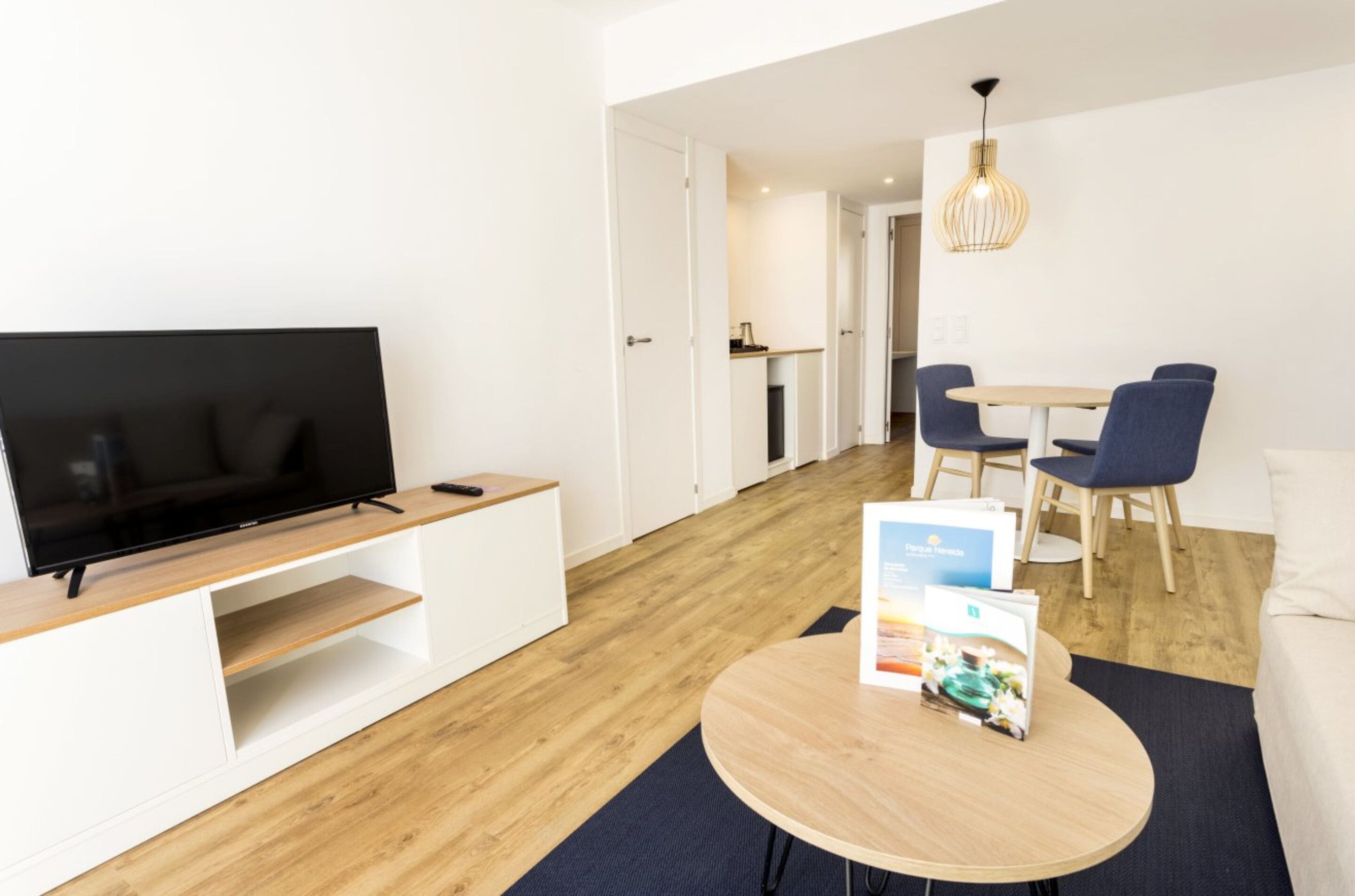 A modern hotel room with a flat-screen TV on a wooden stand, a dining area with a table and chairs, and a hallway leading to a kitchen.