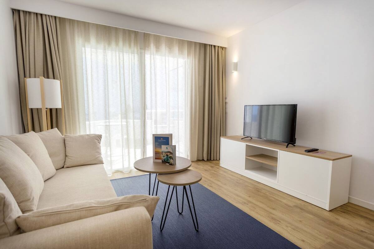 A modern hotel room with a beige sofa, a small round table with a magazine, a TV on a wooden stand, and large windows with sheer curtains.