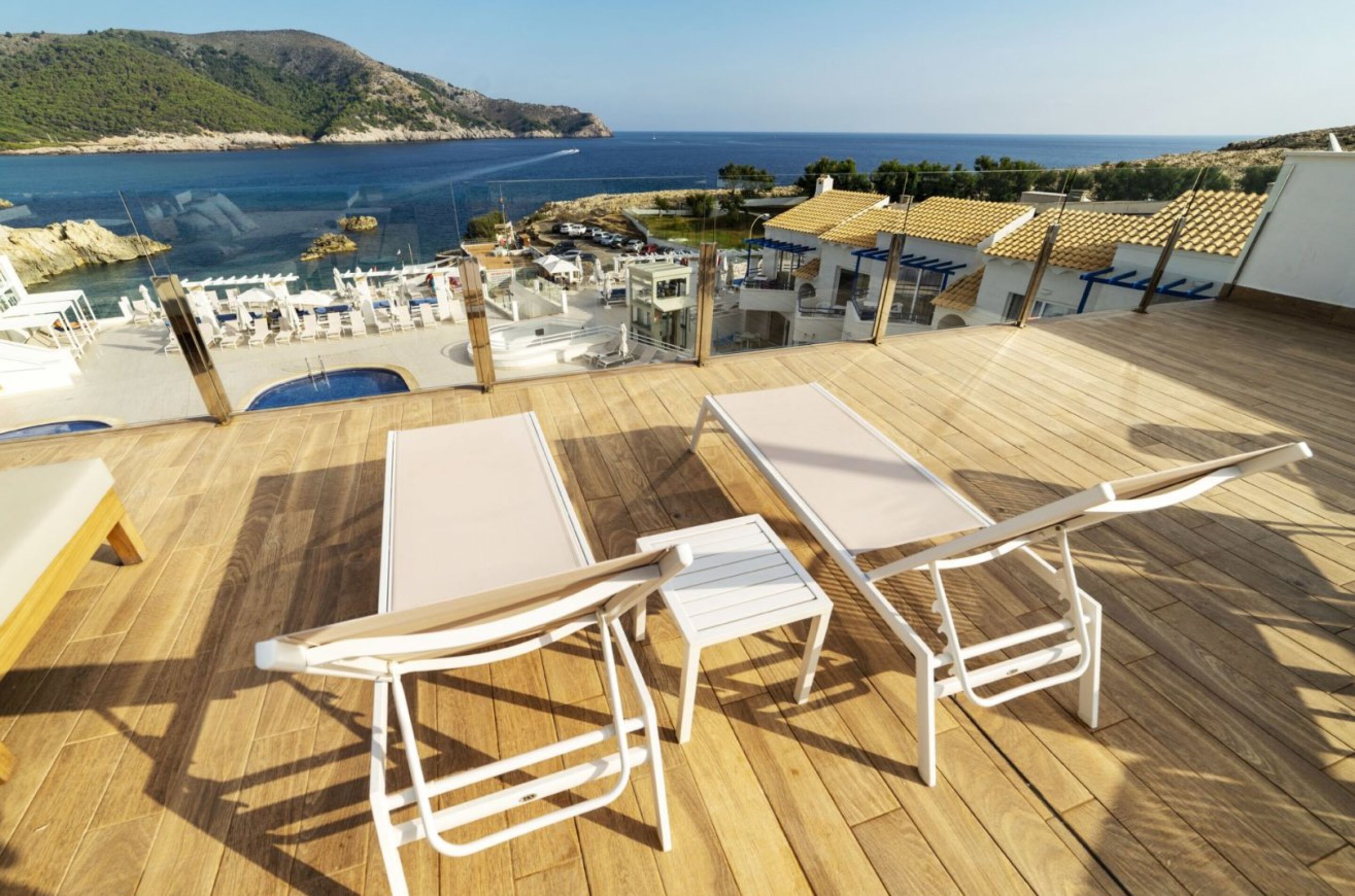 A wooden deck with two white lounge chairs and a table, overlooking a coastal area with buildings and a view of the sea.