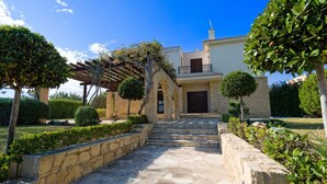 A two-story house with a stone facade, a covered entrance with a pergola, and a well-maintained garden with trimmed bushes and trees. - Villa 64 Anarita - Three Bedroom Villa, Sleeps 6 (Kouklia)