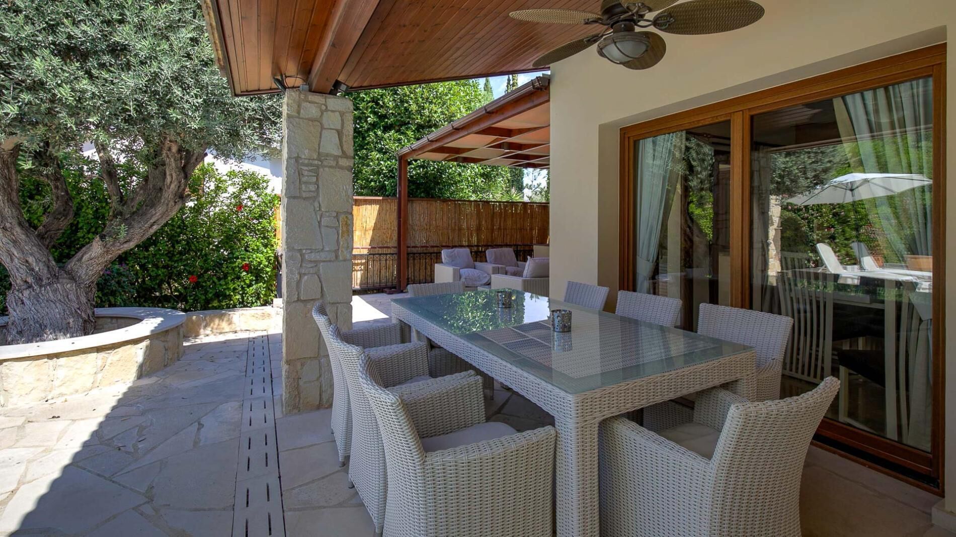 An outdoor patio with a glass-top table and wicker chairs, surrounded by stone walls and a wooden pergola.