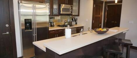 A modern kitchen with dark wood cabinets, stainless steel appliances, a white countertop, and bar stools.