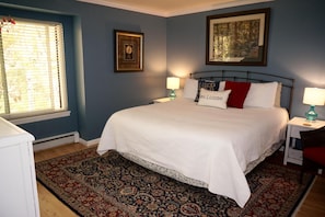 A bedroom with a large bed, two bedside tables with lamps, a patterned rug, and a window with blinds.