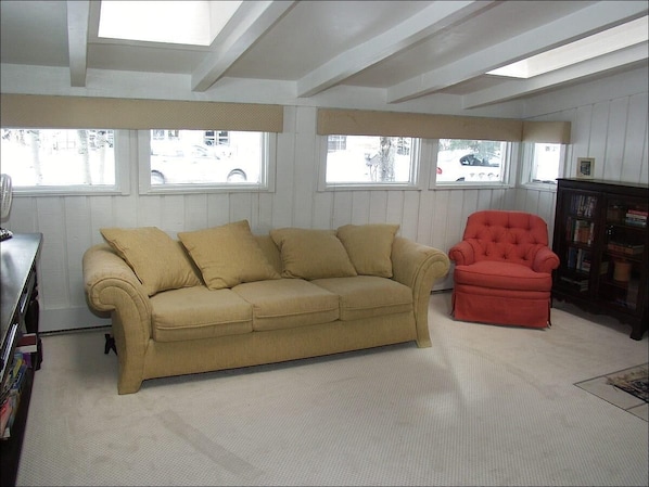 A room with a beige sofa, a red armchair, and a bookshelf. There are windows with blinds, and a ceiling fan is visible. - 733 Francis St W (Aspen)