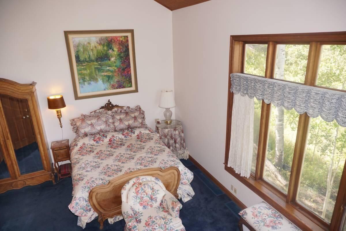 A bedroom with a floral bedspread, a wooden headboard, a bedside table with a lamp, a large window with a view of trees, and a wardrobe.