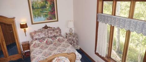 A bedroom with a floral bedspread, a wooden headboard, a bedside table with a lamp, a large window with a view of trees, and a wardrobe.