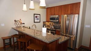A kitchen with granite countertops, wooden bar stools, stainless steel appliances, and pendant lighting.