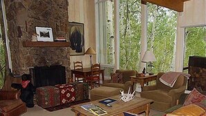 A cozy living room with a stone fireplace, comfortable seating, and large windows offering a view of greenery. - 1131 Faraway Rd (Snowmass Village)