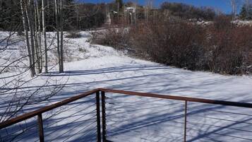 Schneebedecktes Gelände im Freien mit einem Metallgeländer, kahl stehenden Bäumen und einem Gebäude in der Ferne.