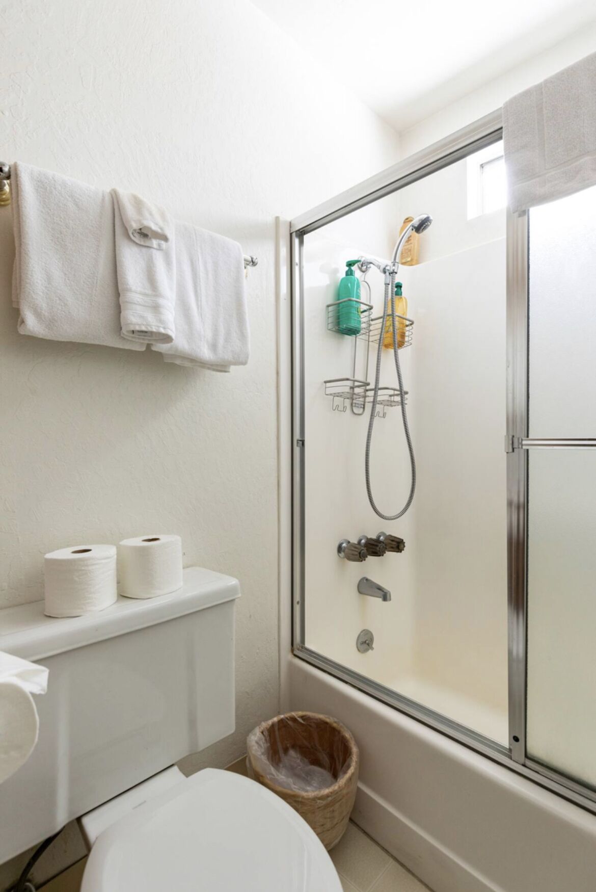 A bathroom with a glass-enclosed shower, toilet paper, and a waste bin.