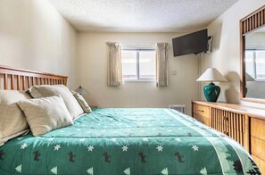 Ein Schlafzimmer mit einem Bett, einem an der Wand montierten Fernseher, einer Lampe und einem Fenster mit Vorhängen.
