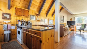 A cozy cabin interior with wooden walls, a kitchen with a central island, a living area with a sofa and TV, and a large window offering a view.