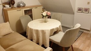 Ein gemütlicher Raum mit einem runden Tisch, zwei Stühlen, einer Couch, einem Schrank mit einem Radio und einer Vase mit Blumen.