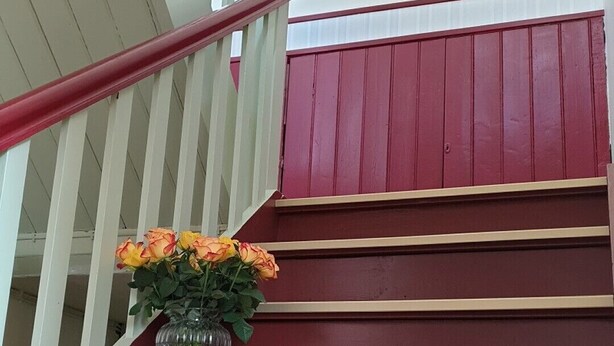 Eine Treppe mit roten Stufen und weißen Geländern, auf der ersten Stufe ein Blumenvasen mit gelben Blumen.