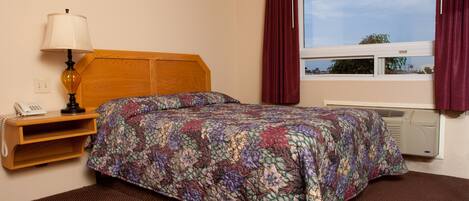 A hotel room with a wooden headboard, a floral bedspread, a bedside lamp, a telephone, and a window with curtains.