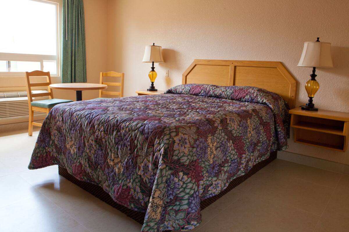 Quarto de hotel com uma cama grande, duas mesas de cabeceira com abajures, uma cadeira e uma mesinha.