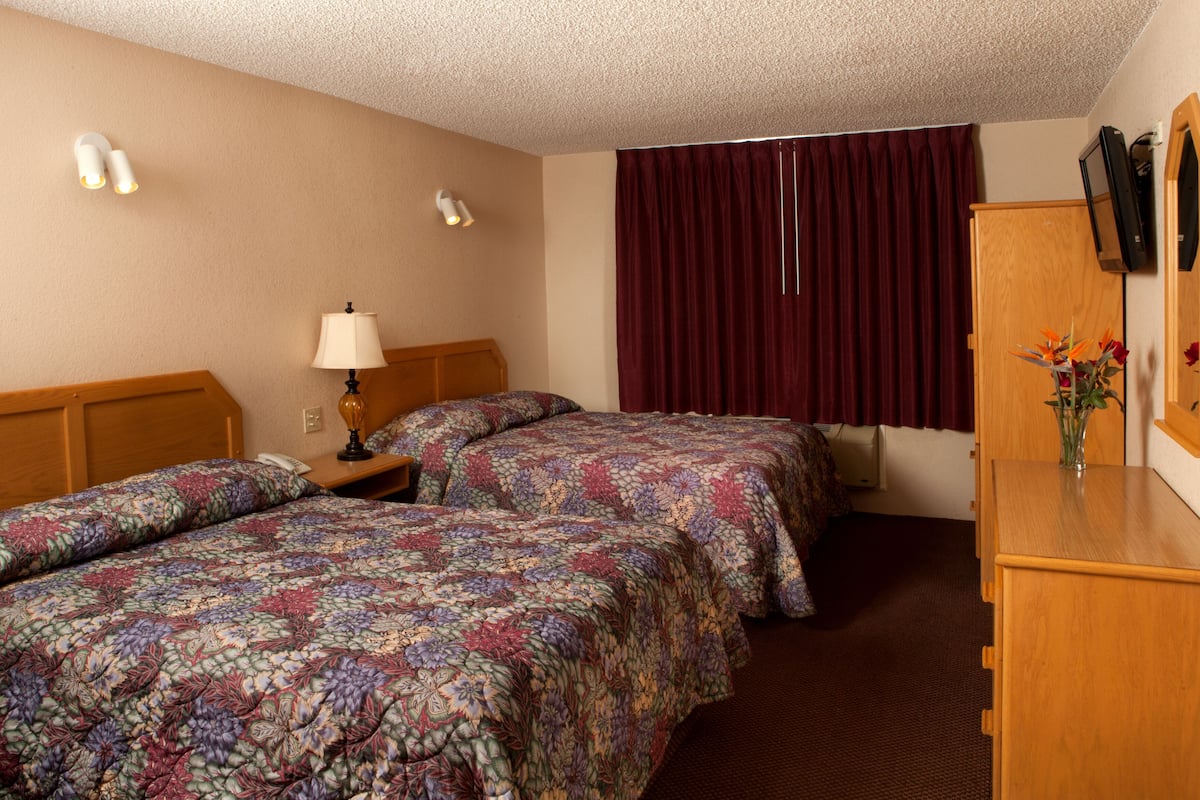 Quarto de hotel com duas camas, uma mesa de cabeceira, abajur, espelho, cômoda e janela com cortinas.