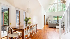 Un vaste espace dédié à la salle à manger, avec une table en bois et des chaises blanches, un luminaire suspendu et une vue sur l’extérieur grâce à de grandes fenêtres.