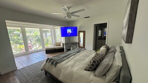 Camera da letto con un letto, una ventola a soffitto, una TV e vista su del verde attraverso ampie finestre.