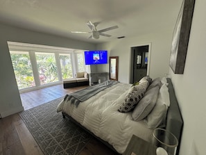 Ein Schlafzimmer mit einem Bett, einem Deckenventilator, einem Fernseher und Blick auf Grün hinter großen Fenstern.