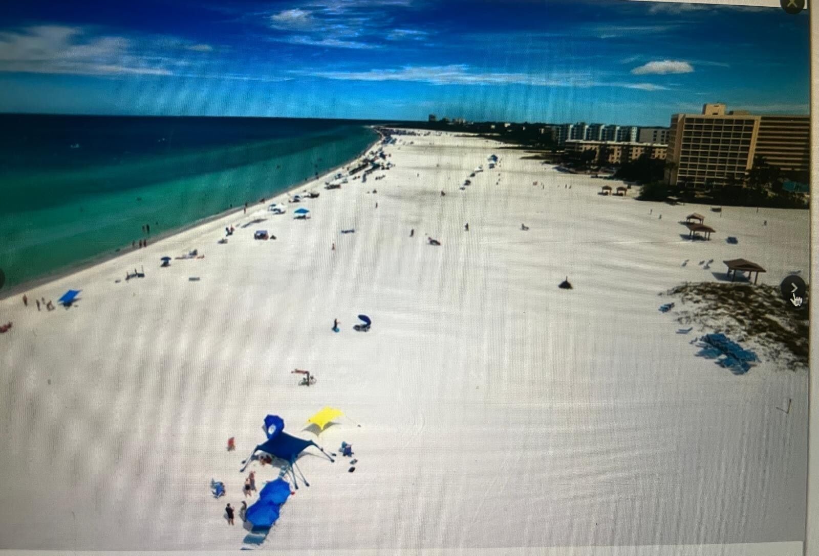 Ein Strand mit blauem Himmel, sandigem Ufer und Gebäuden im Hintergrund.