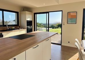 Een moderne keuken met een houten aanrecht, witte kasten en uitzicht op de omgeving door grote glazen schuifdeuren.