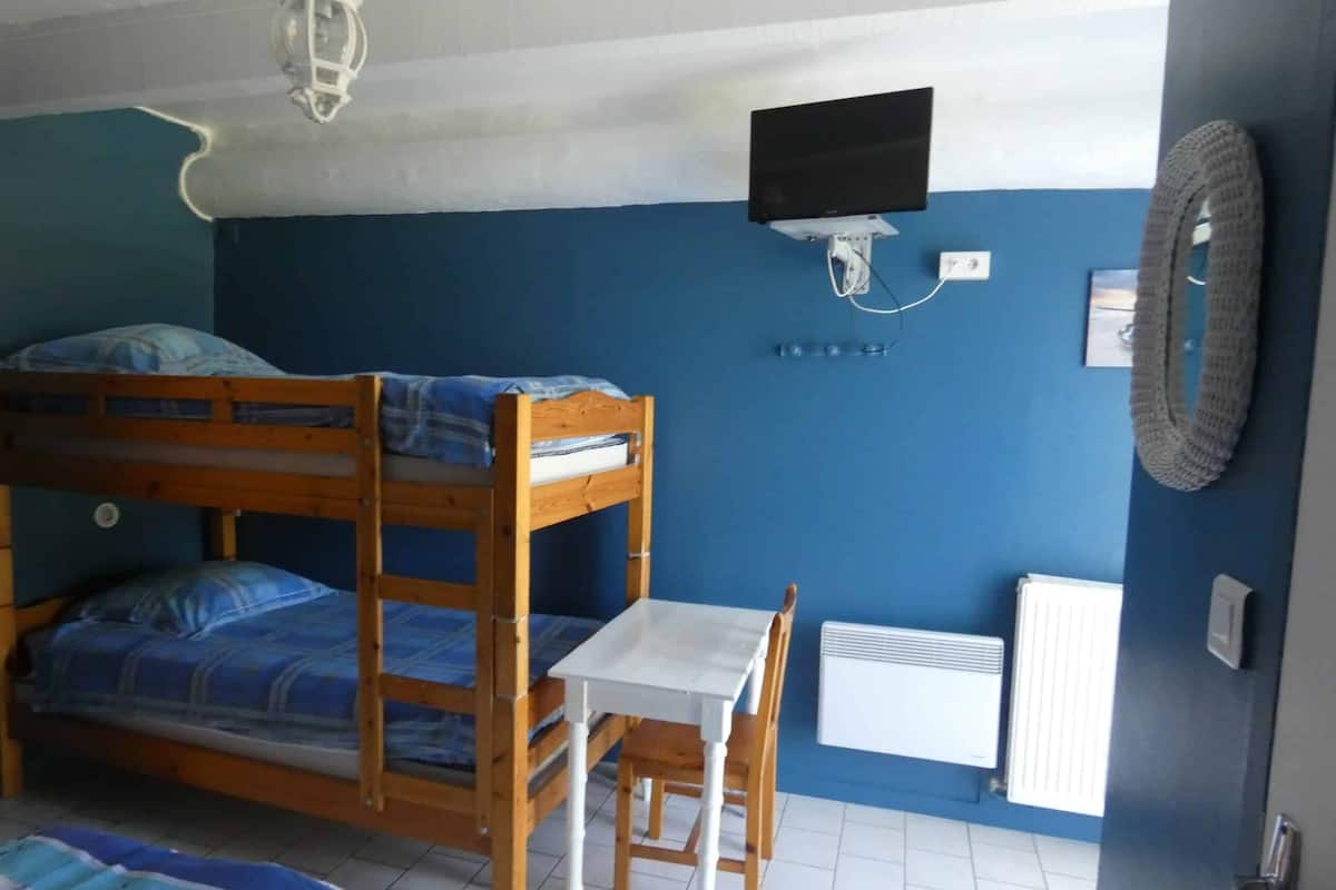 A room with a bunk bed, a small table, a chair, a TV mounted on the wall, and a radiator.