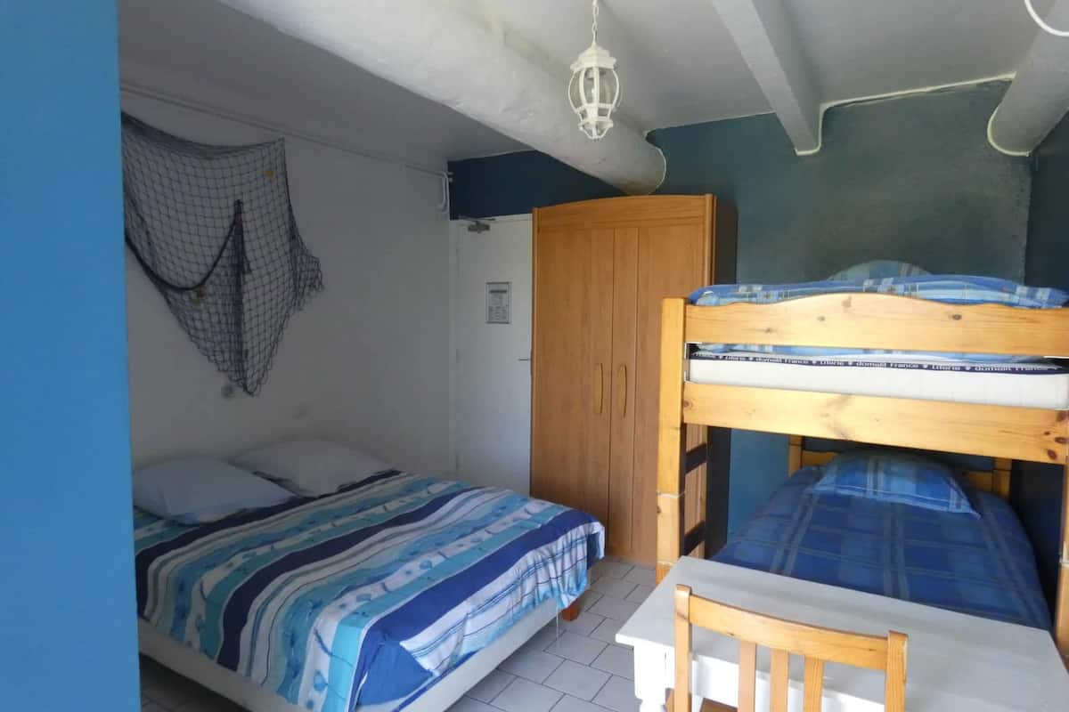 A small, compact room with a bunk bed, a single bed, a wooden table, and a hanging light fixture.