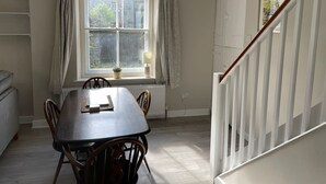 A dining area with a table and chairs, a window with curtains, a radiator, and a staircase with a white railing. - Bluebell Cottage, Characterful Cottage in the Yorkshire Dales (Langcliffe)
