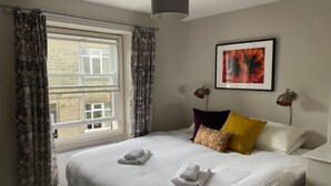A neatly made bed with white linens and two towels, a window with patterned curtains, and a framed abstract painting on the wall. - Bluebell Cottage, Characterful Cottage in the Yorkshire Dales (Langcliffe)