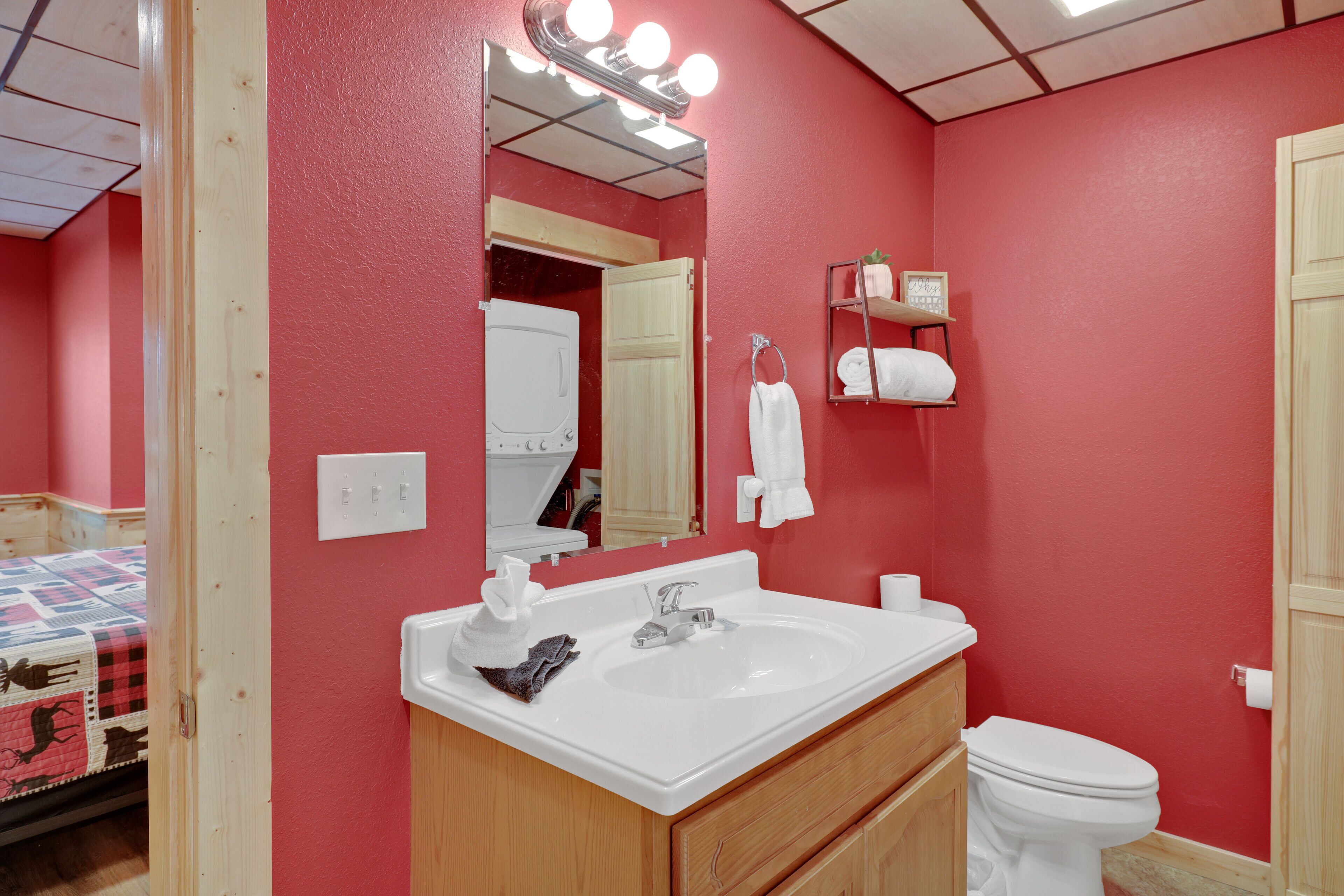 A bathroom with a red wall, a white sink, a mirror with lights, a white toilet, and a wooden cabinet.