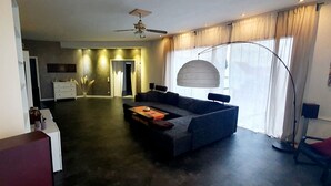 Una sala de estar moderna con un sofá gris oscuro, una mesa de centro de madera y un ventilador de techo. Hay un ventanal con cortinas translúcidas y una lámpara de pie.