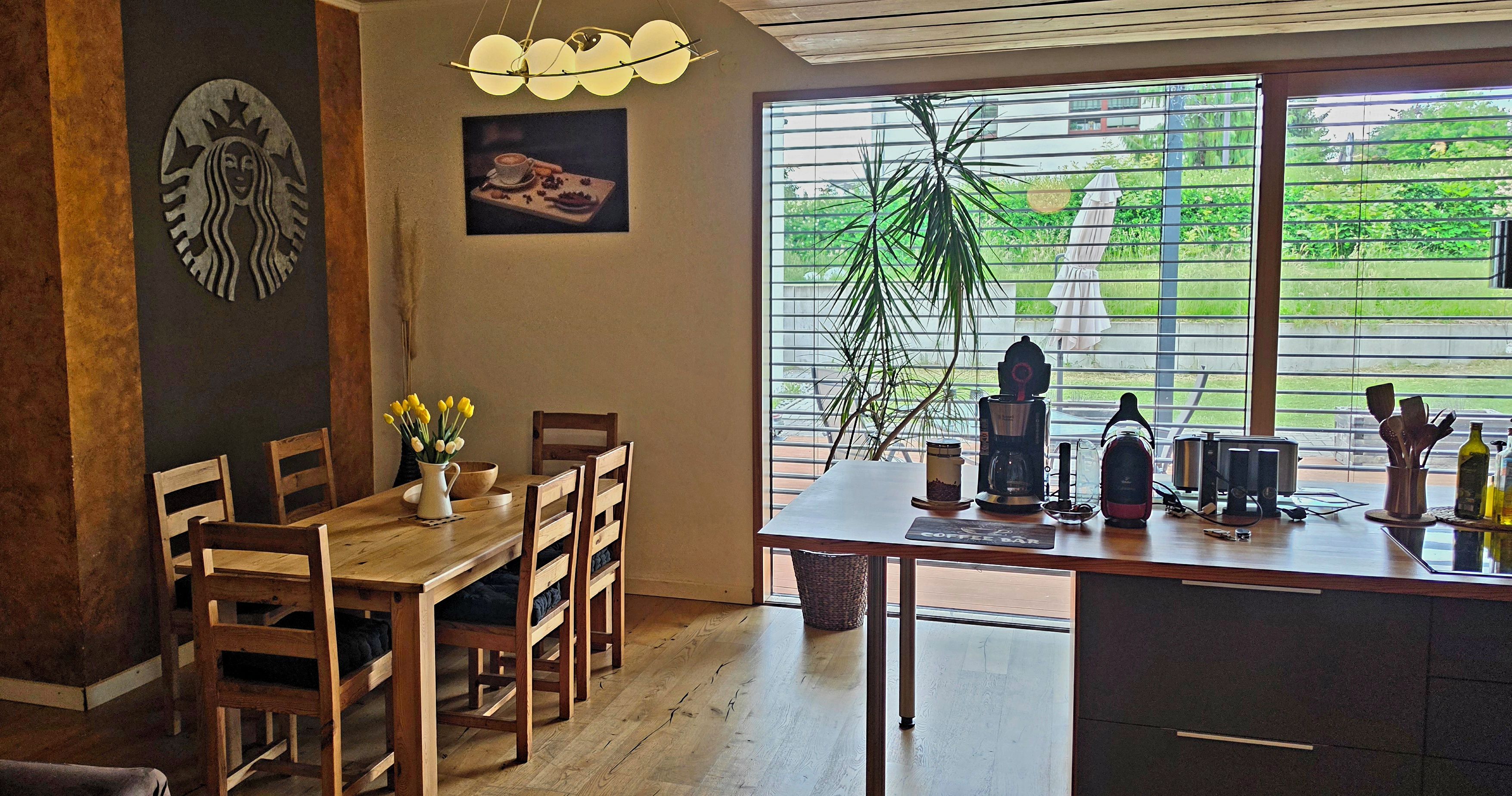 A dining area with a wooden table, chairs, a vase of yellow flowers, a coffee machine, and a view of greenery through large windows.