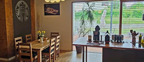 A dining area with a wooden table, chairs, a vase of yellow flowers, a coffee machine, and a view of greenery through large windows.