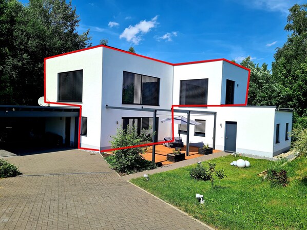 Una casa moderna de dos pisos con fachada blanca y detalles rojos, que cuenta con un porche cubierto y mobiliario de exterior.