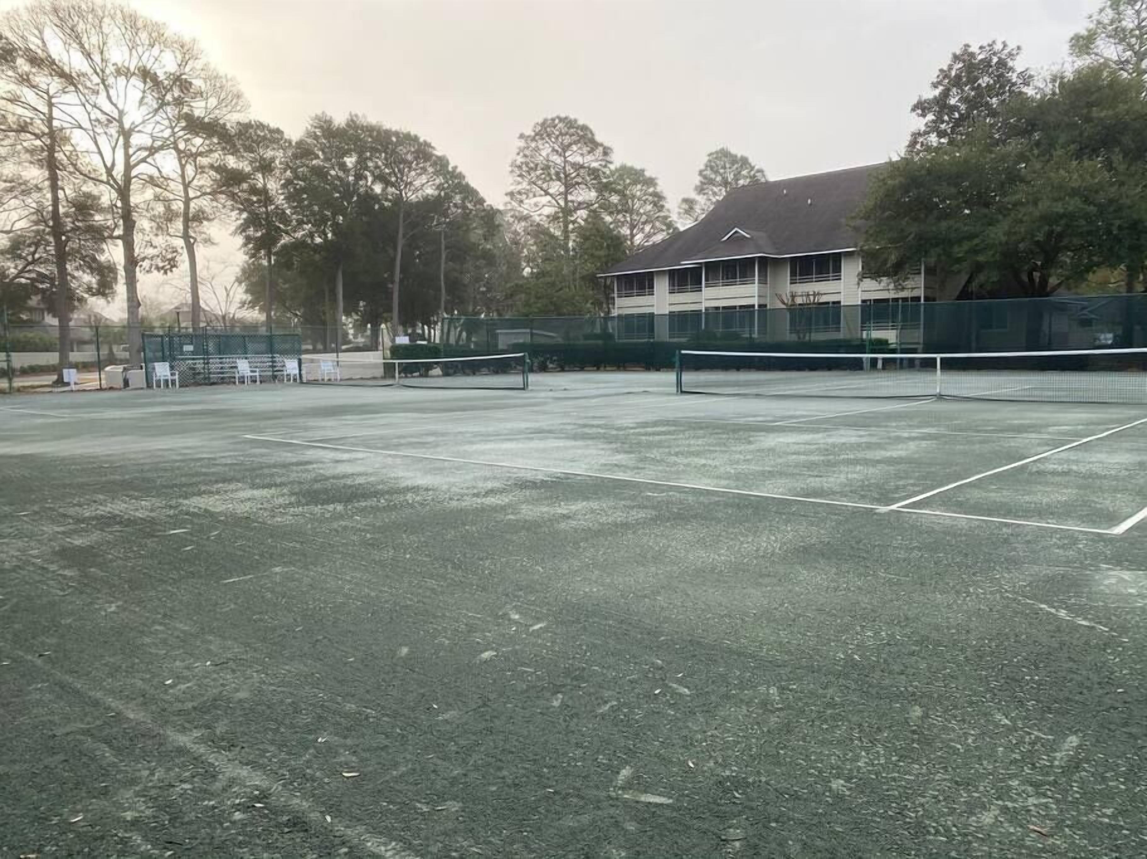An outdoor tennis court with a net, surrounded by trees and a building in the background.