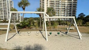 A playground with swings and a sandbox in a park area with palm trees and a building in the background.