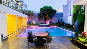 Una piscina con una mesa y sillas de madera en el centro, rodeada por un edificio moderno con un muro verde y un cartel que dice "RAM".