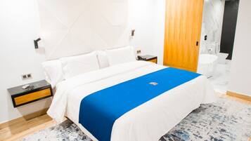 Una habitación de hotel moderna con una cama bien hecha, una mesita de noche con lámpara y un baño a la vista a través de una puerta abierta.