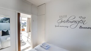 Ein Schlafzimmer mit einem Bett, einer Wand mit dem Schriftzug 'Bonjour Guten Morgen Buenos' und Blick ins Freie.