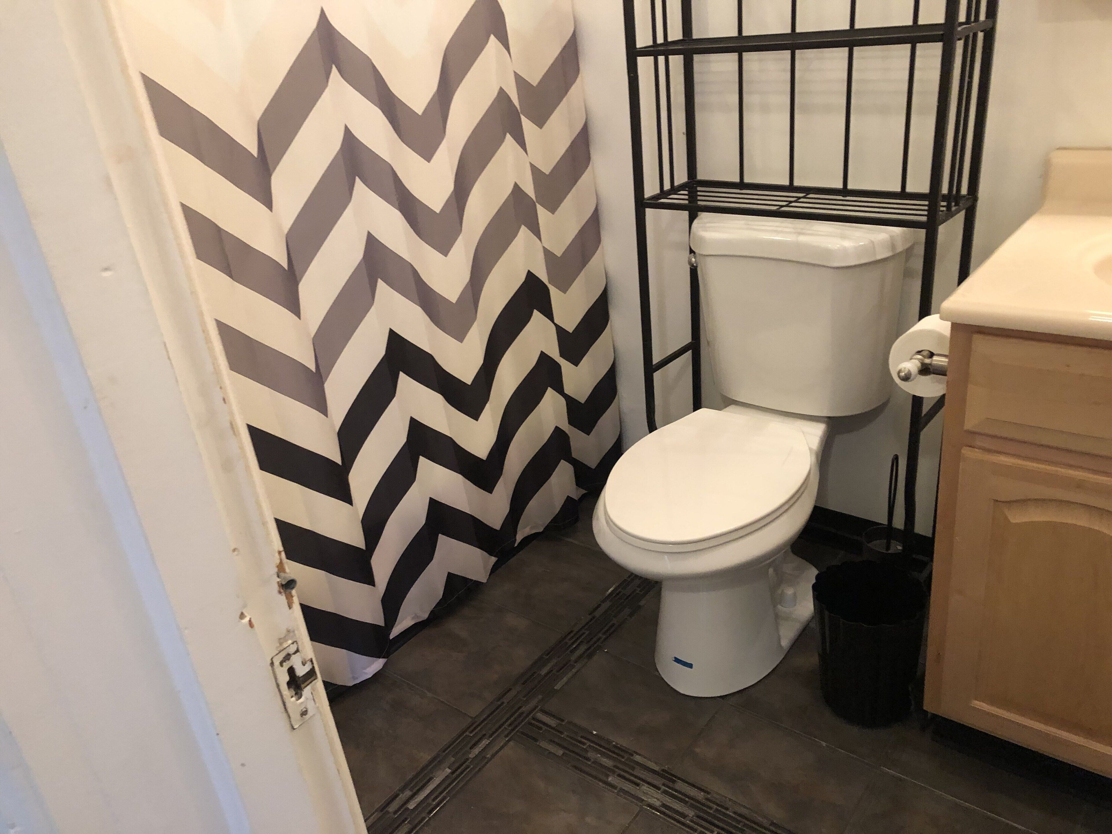 Ein Badezimmer mit einer weißen Toilette, einem schwarz-weißen Duschvorhang mit Zickzackmuster, einem schwarzen Metallregal und einem Holzschrank.