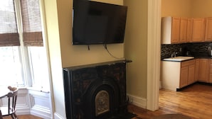 A living room with a fireplace, a television mounted above it, wooden flooring, a dining table, and a kitchen area with wooden cabinets. - 1bedroom/1 bath      (Oneonta)