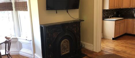 A living room with a fireplace, a television mounted above it, wooden flooring, a dining table, and a kitchen area with wooden cabinets.