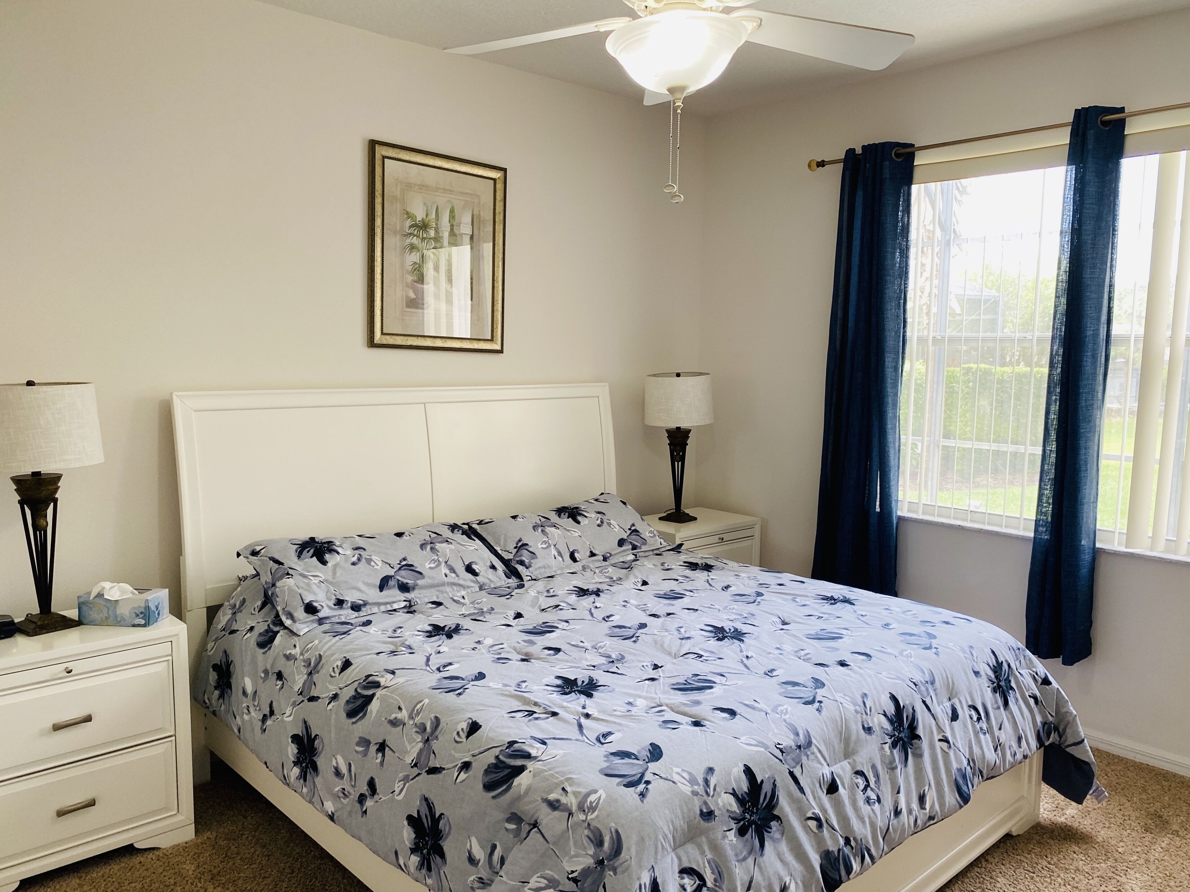 Une chambre à coucher comprenant un lit, deux tables de chevet, un ventilateur de plafond, une fenêtre avec des rideaux et un tableau encadré accroché au mur.