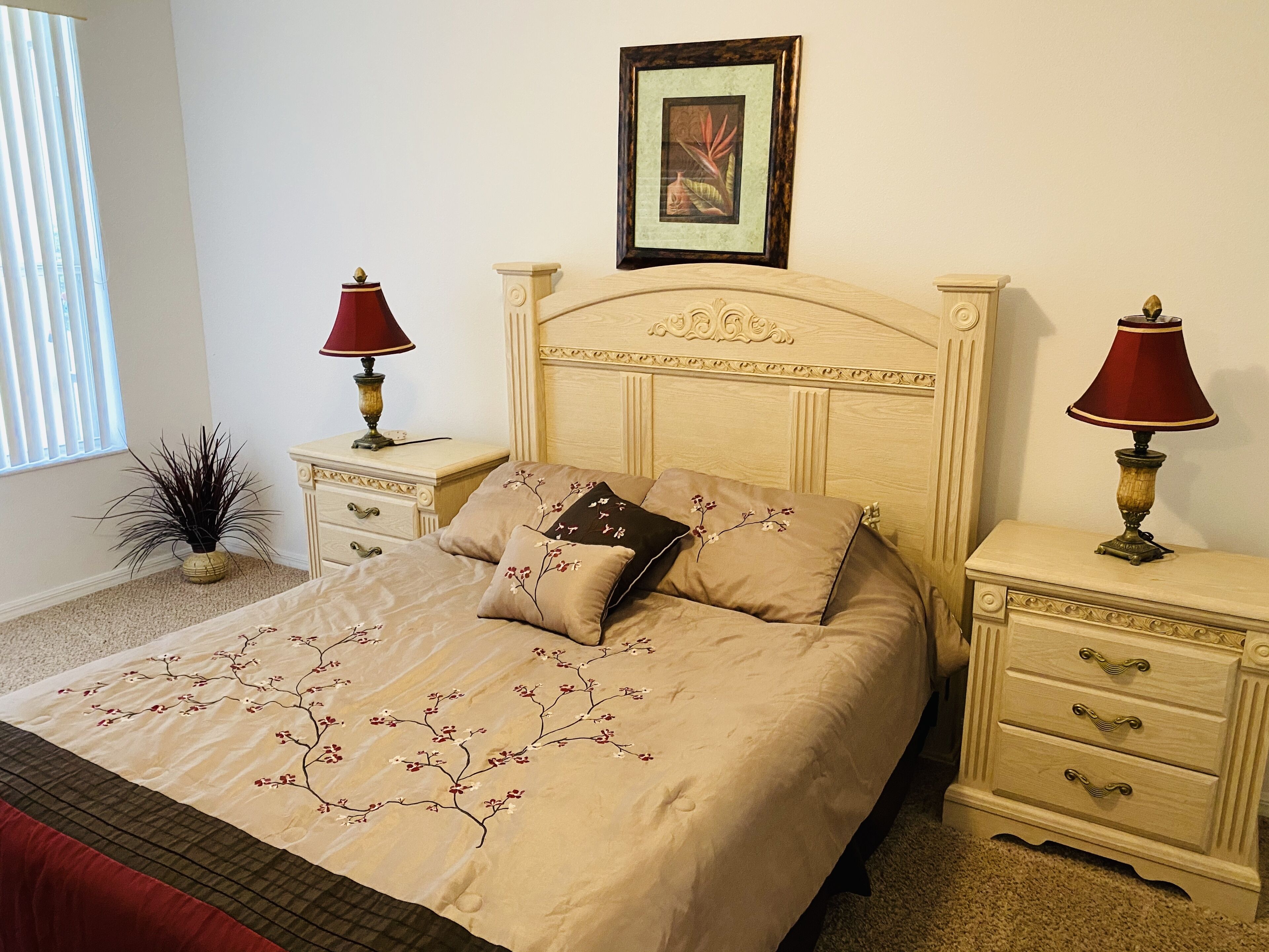 A neatly made bed with floral bedding, two bedside tables with lamps, a framed picture above the headboard, and a potted plant beside the window.