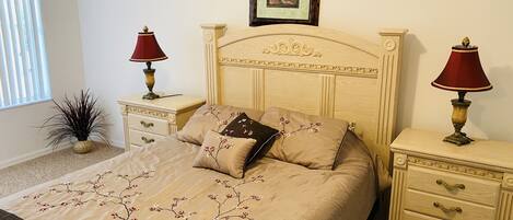 A neatly made bed with floral bedding, two bedside tables with lamps, a framed picture above the headboard, and a potted plant beside the window.