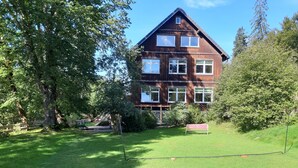 A two-story wooden house with a sloped roof, surrounded by lush greenery and trees. - dasokerhaus.com (Altenau)