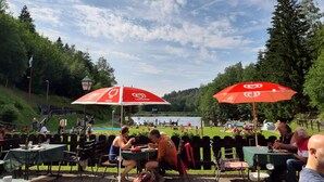 An outdoor dining area with tables and chairs under red umbrellas, set against a backdrop of a lake and trees. - dasokerhaus.com (Altenau)
