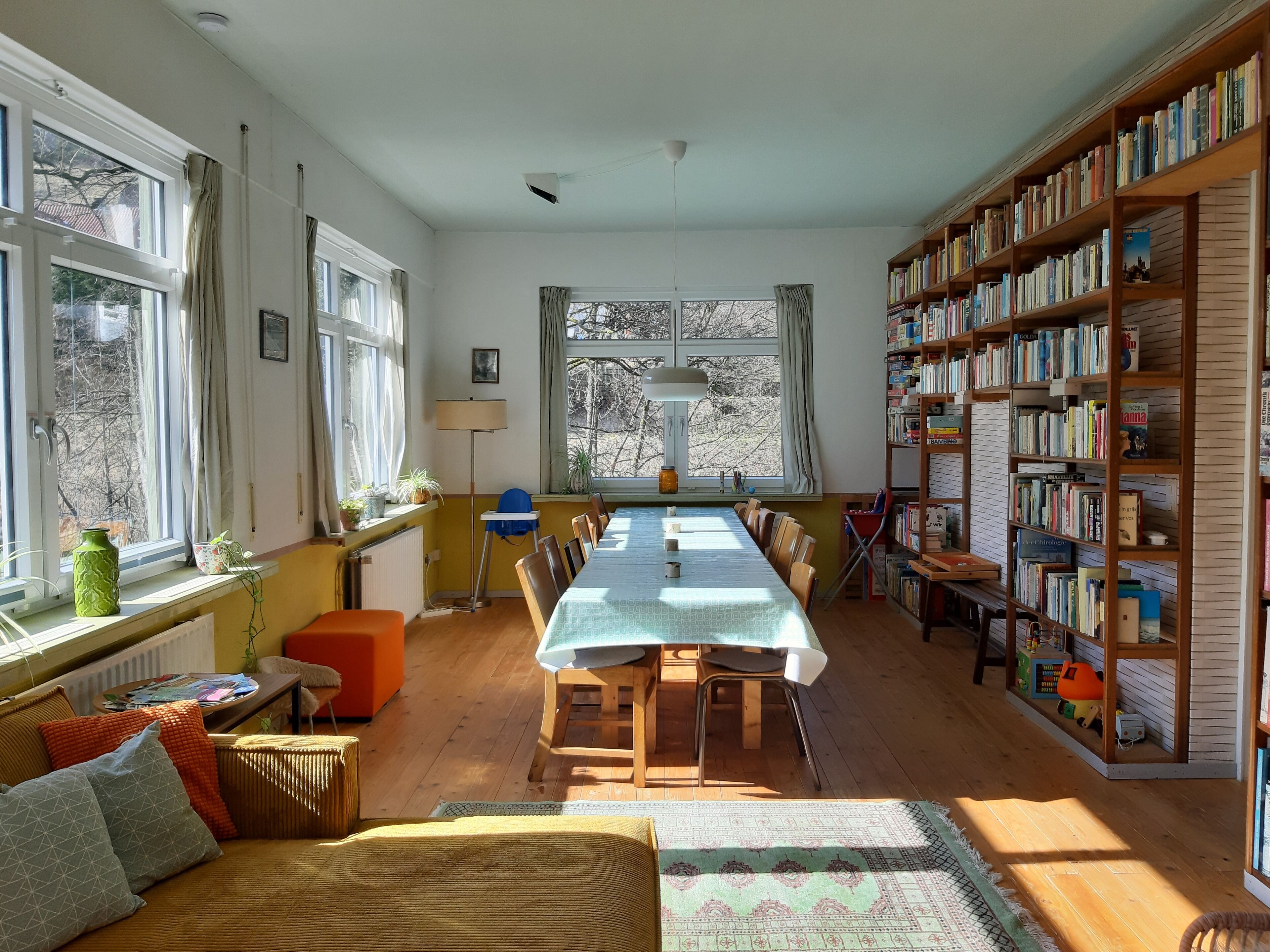 Um quarto acolhedor com uma mesa comprida, cadeiras, uma estante e uma janela com cortinas.