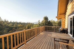 Une terrasse en bois avec une rambarde, une table et un barbecue, donnant sur une zone boisée.